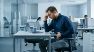 Toxic Workplace 2 - Dr Soumya Dash Man in blue shirt sits at a desk in an office, head in hand, looking stressed while working on a laptop.
